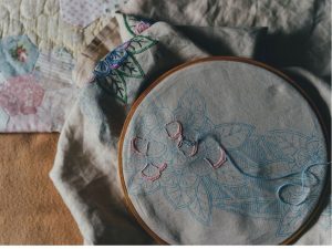 Embroidery hoop with embroidered floral design next to antique quilt.
